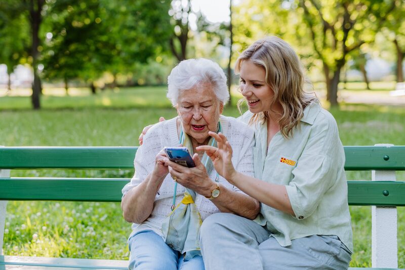 Adult granddaguhter helping her grandmother to use cellphone when sitting on bench in park in summer.