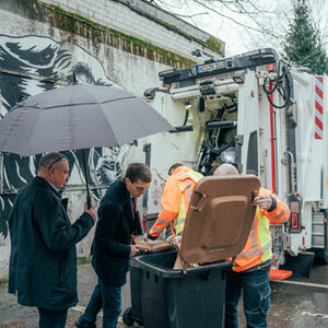 Männer schauen sich Biomülltonne an im Hintergrund ein Müllfahrzeug mit Sensorik
